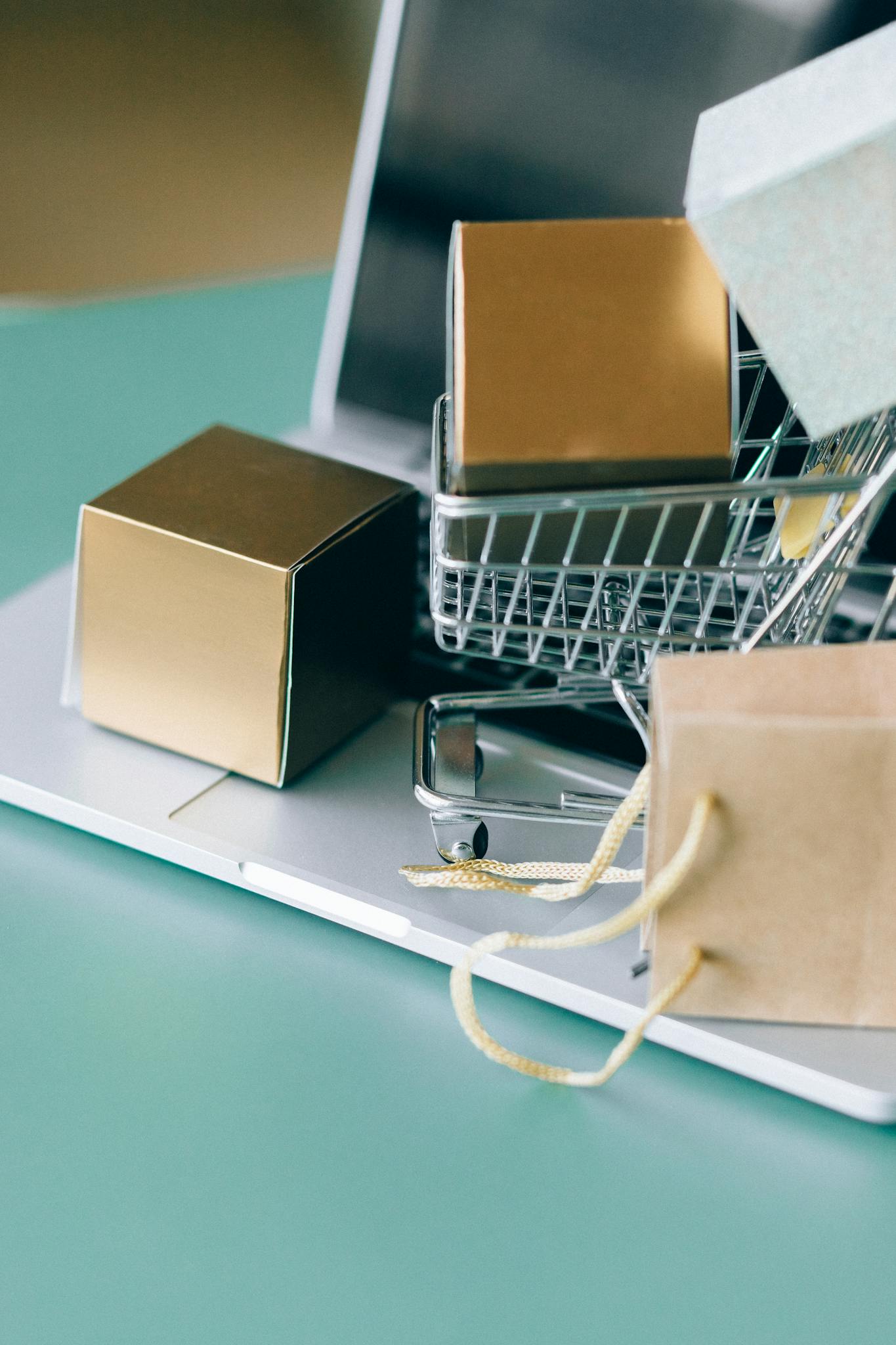 Stylish still life featuring a miniature shopping cart on a laptop with elegant gift boxes, depicting modern e-commerce.
